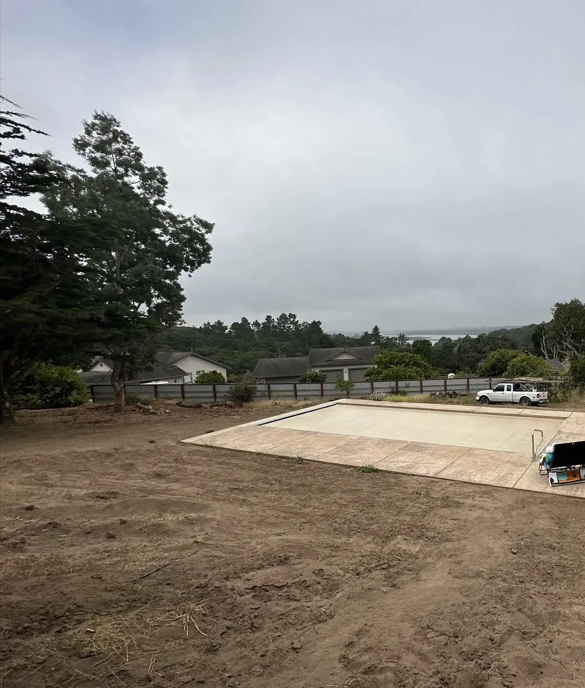 A rectangular swimming pool with a closed cover and stamped concrete decking surrounded by a bare dirt yard.