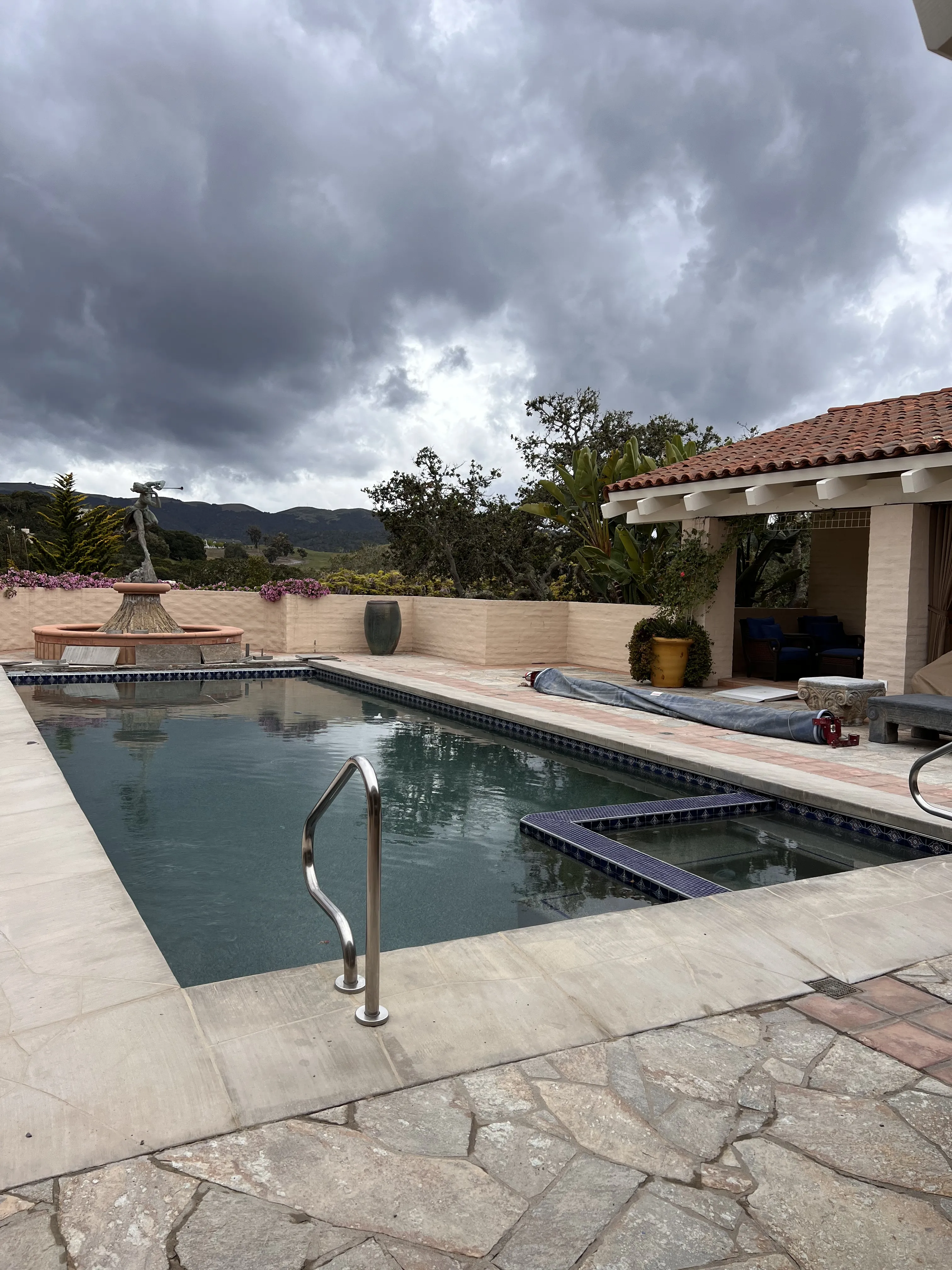 A finished rectangular pool and attached spa with blue tile spillover, surrounded by a flagstone deck under a stormy sky.