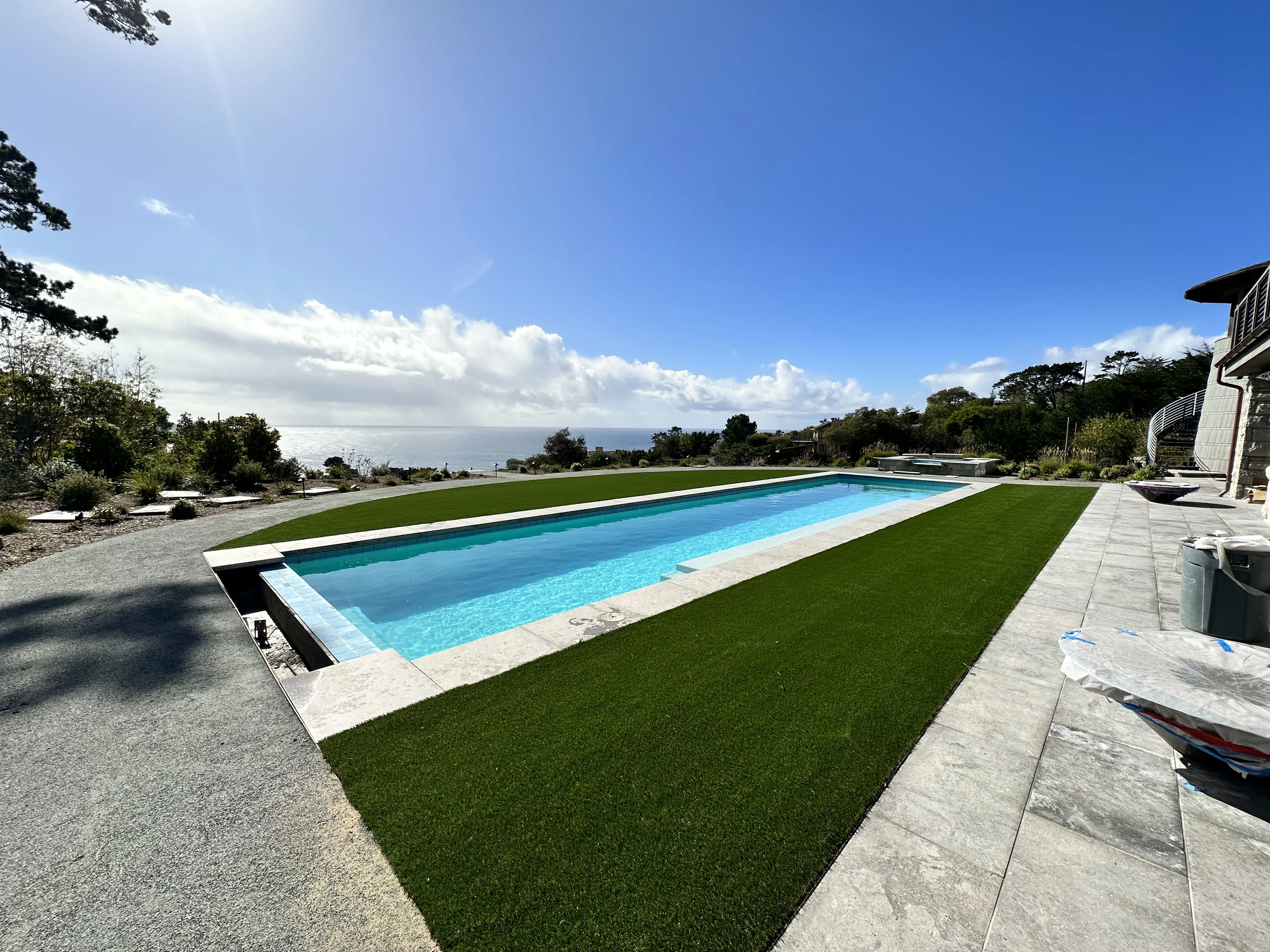 Finished rectangular infinity edge swimming pool surrounded by artificial turf and stone patio.