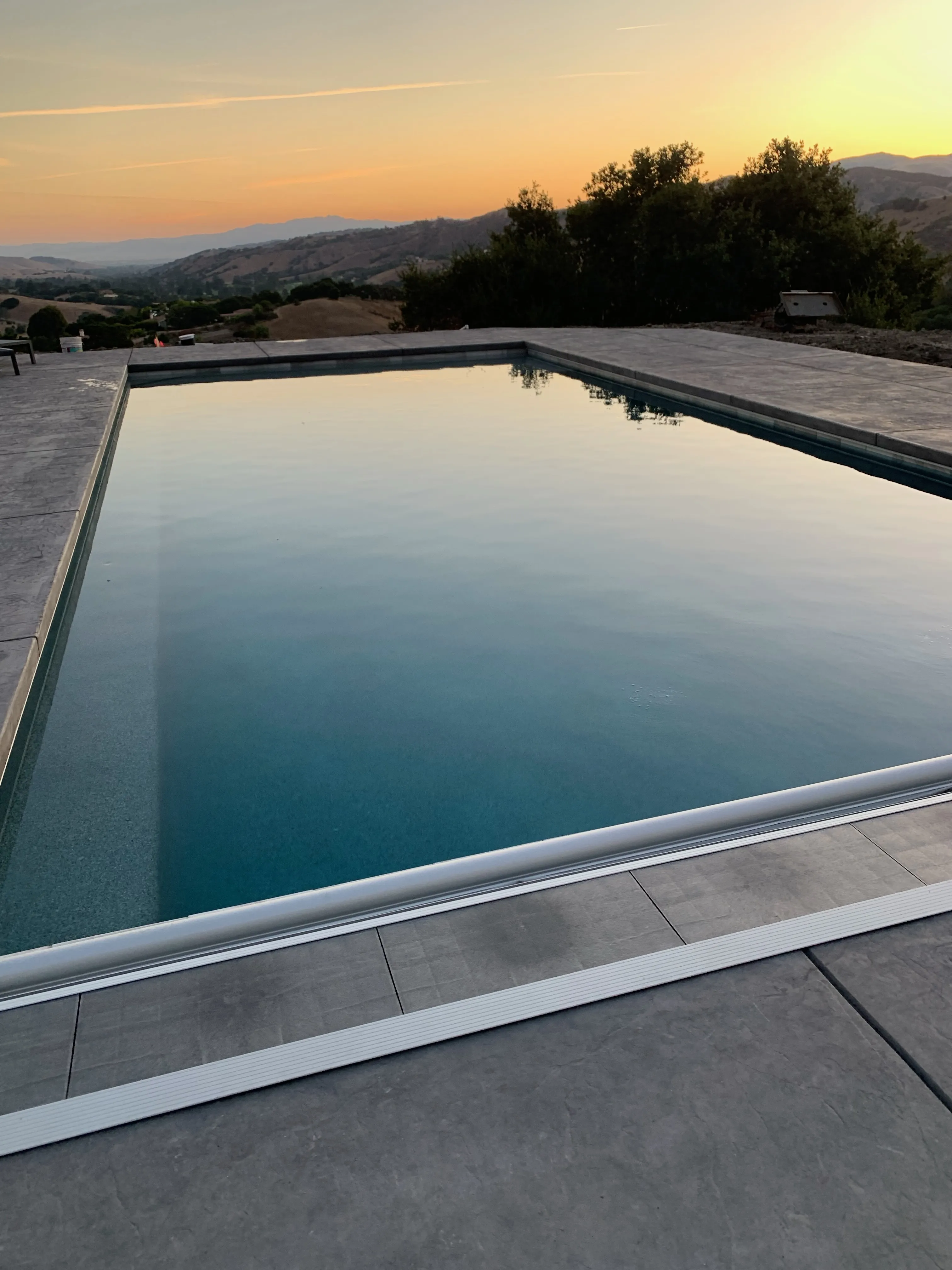 A finished rectangular infinity pool with a concrete deck and automatic cover track overlooking a valley at sunset.