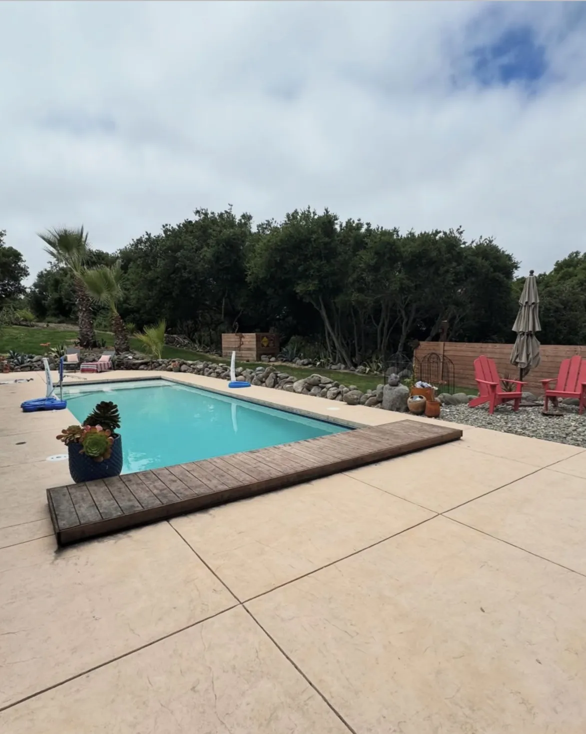 Finished rectangular swimming pool with a wooden platform on a light concrete deck, a rock border, and red chairs.