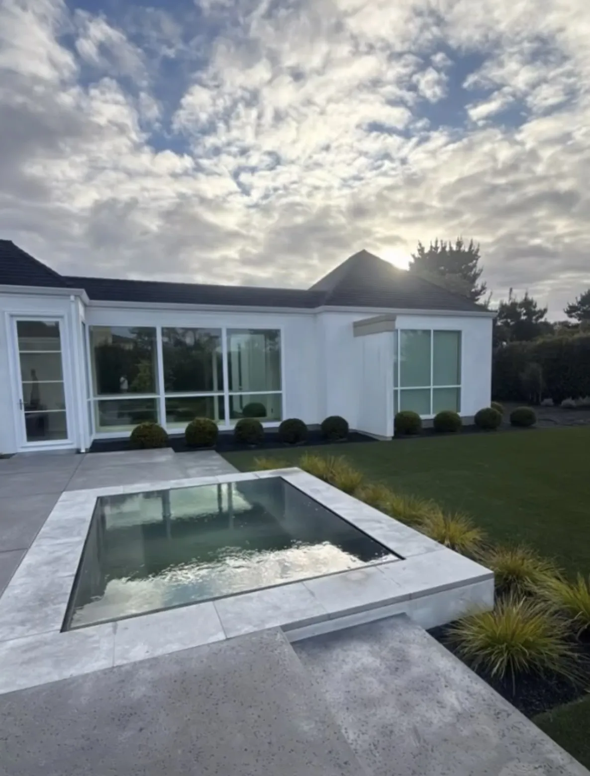 A finished square spa with light grey stone coping in a backyard with a white modern house in the background.