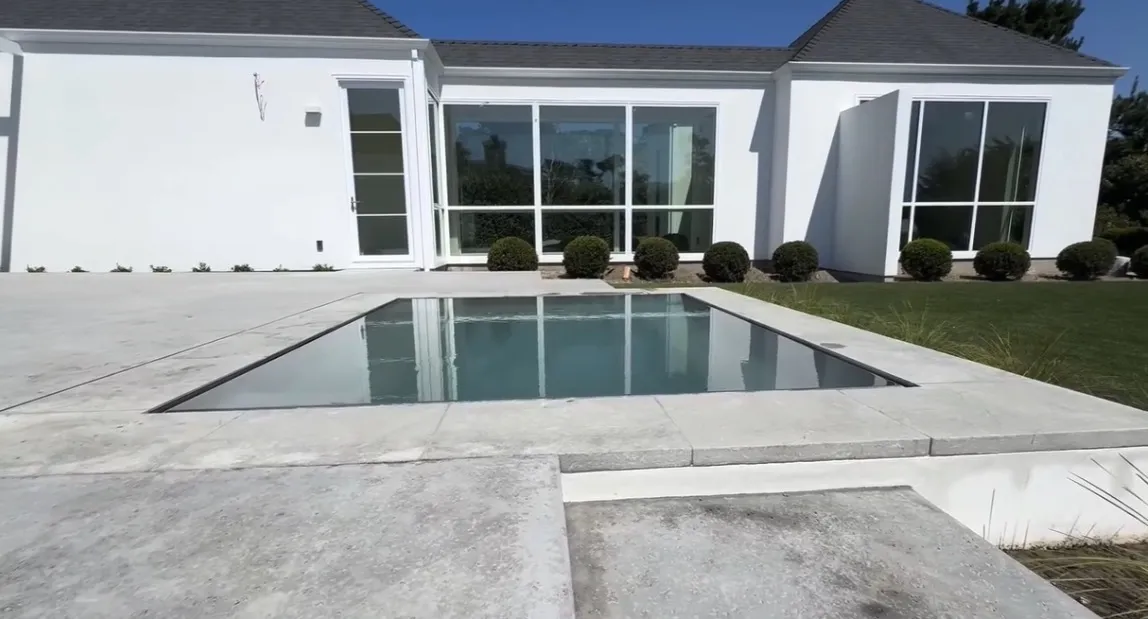 Finished rectangular pool flush with a light grey paved deck, reflecting a white house with a dark pitched roof.