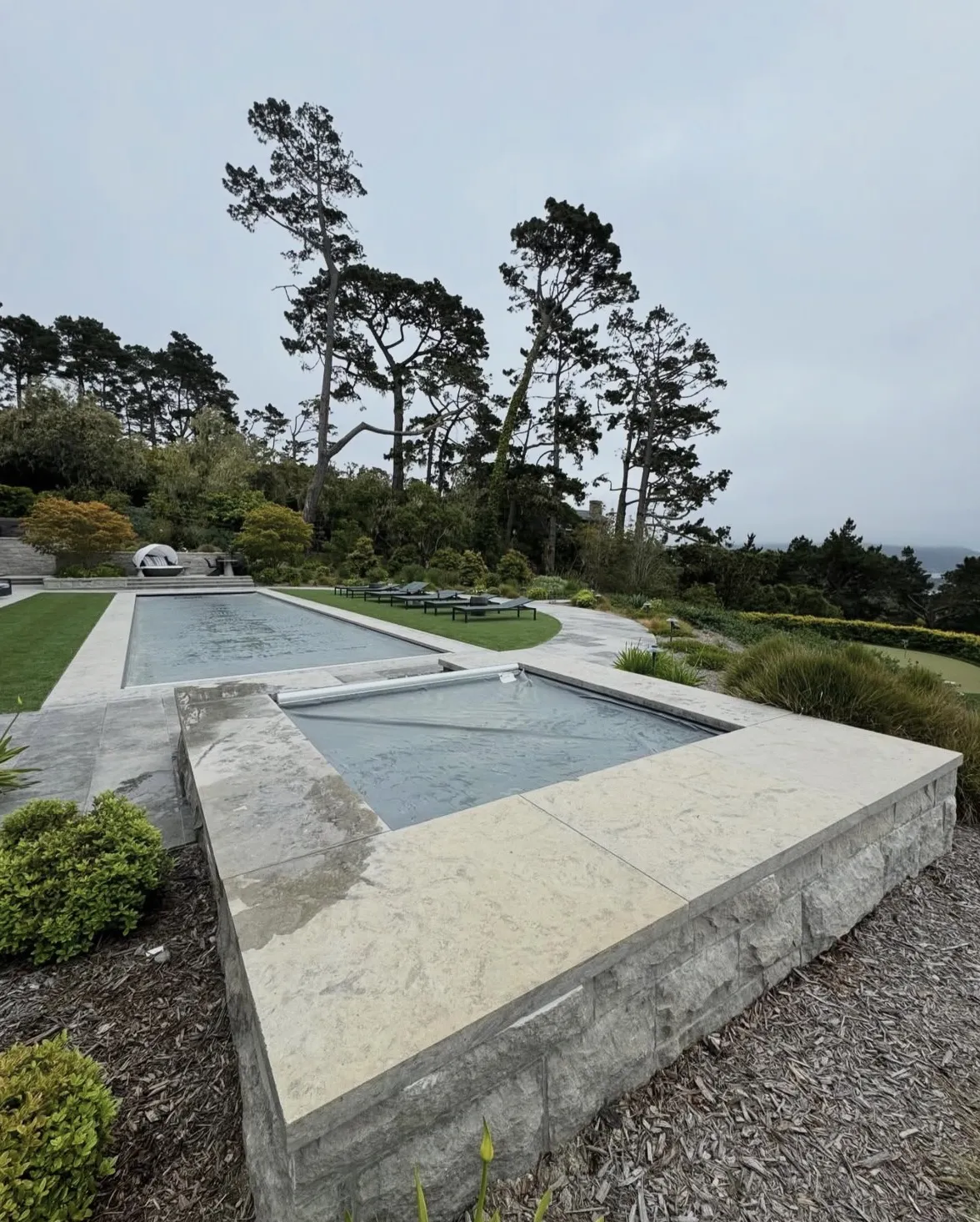 Completed rectangular pool and raised spa with closed automatic covers in a stone patio with lawn and trees.