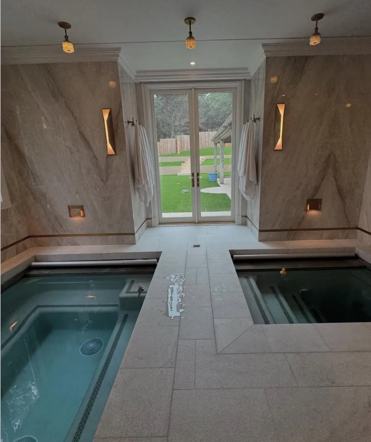 Two indoor spas separated by a light stone walkway in a room with veined walls and glass doors opening to a yard.
