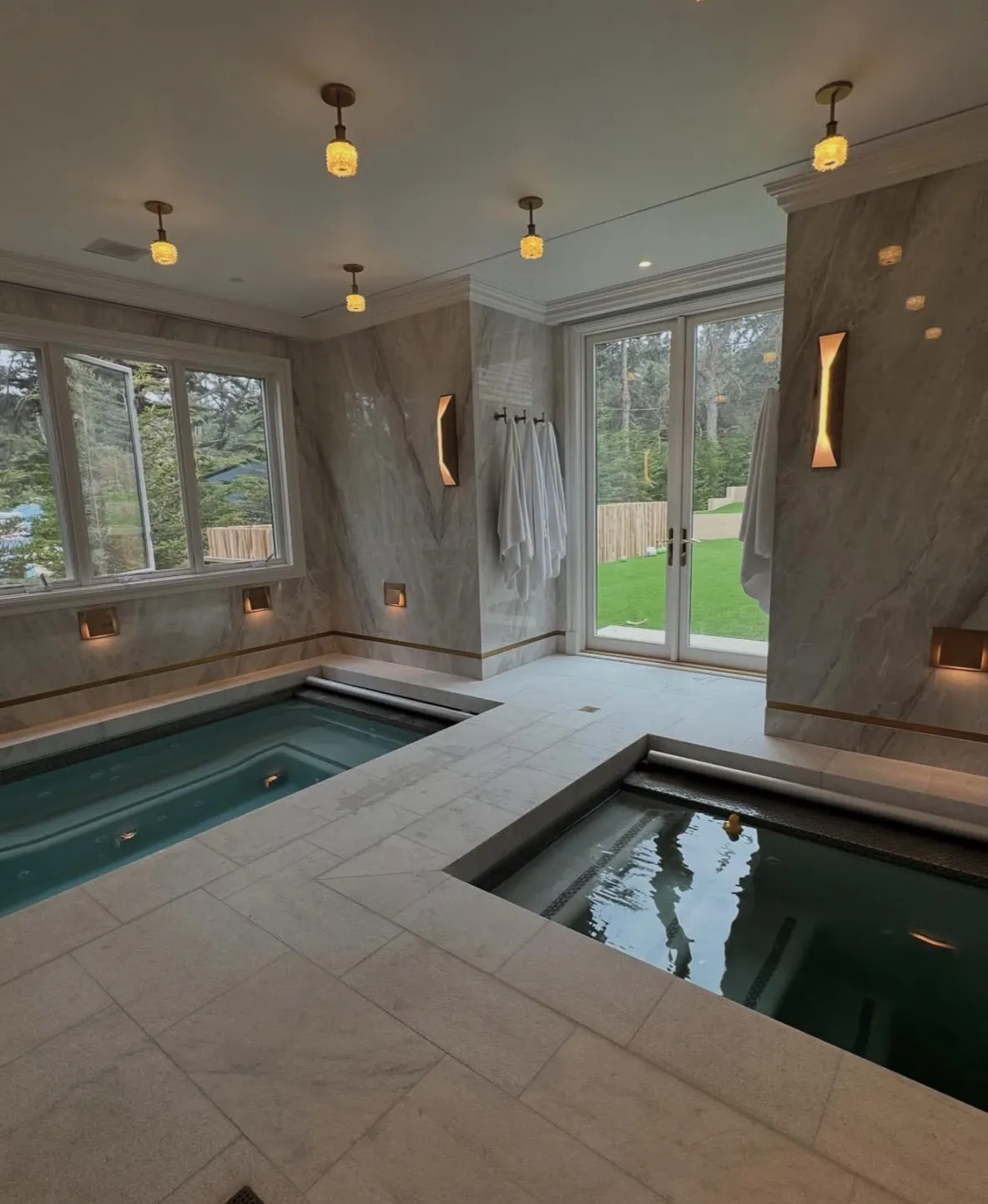 Indoor rectangular pool and spa set into a stone floor inside a marble-walled room with French doors opening to a yard.
