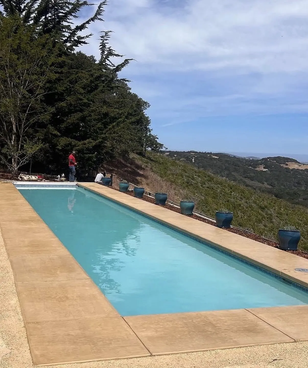 Rectangular lap pool with a tan concrete deck and blue planters overlooking rolling hills.