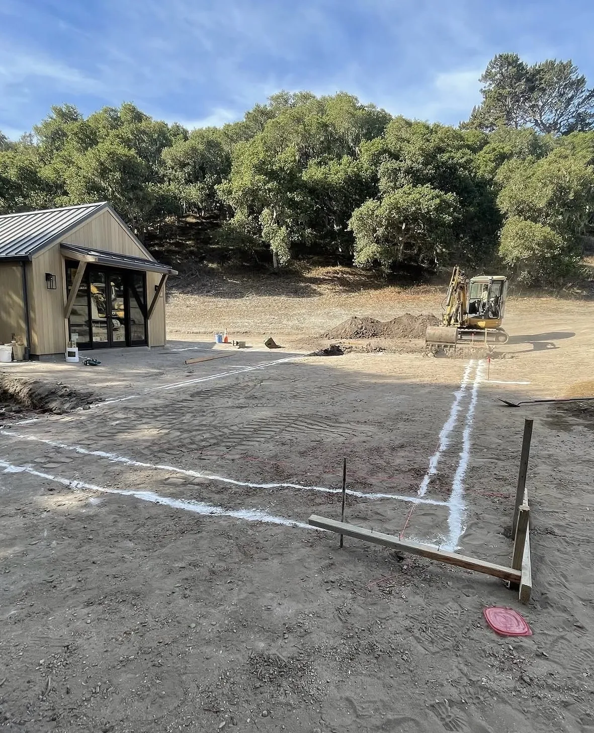 White painted layout lines and wooden stakes mark a pool shape on a dirt site, with a yellow excavator in the background.