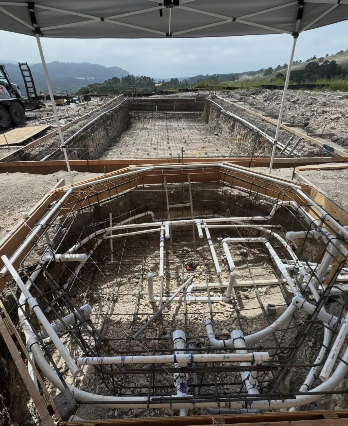 Excavated octagonal spa showing PVC plumbing and rebar, with a rectangular pool's rebar cage in the background.