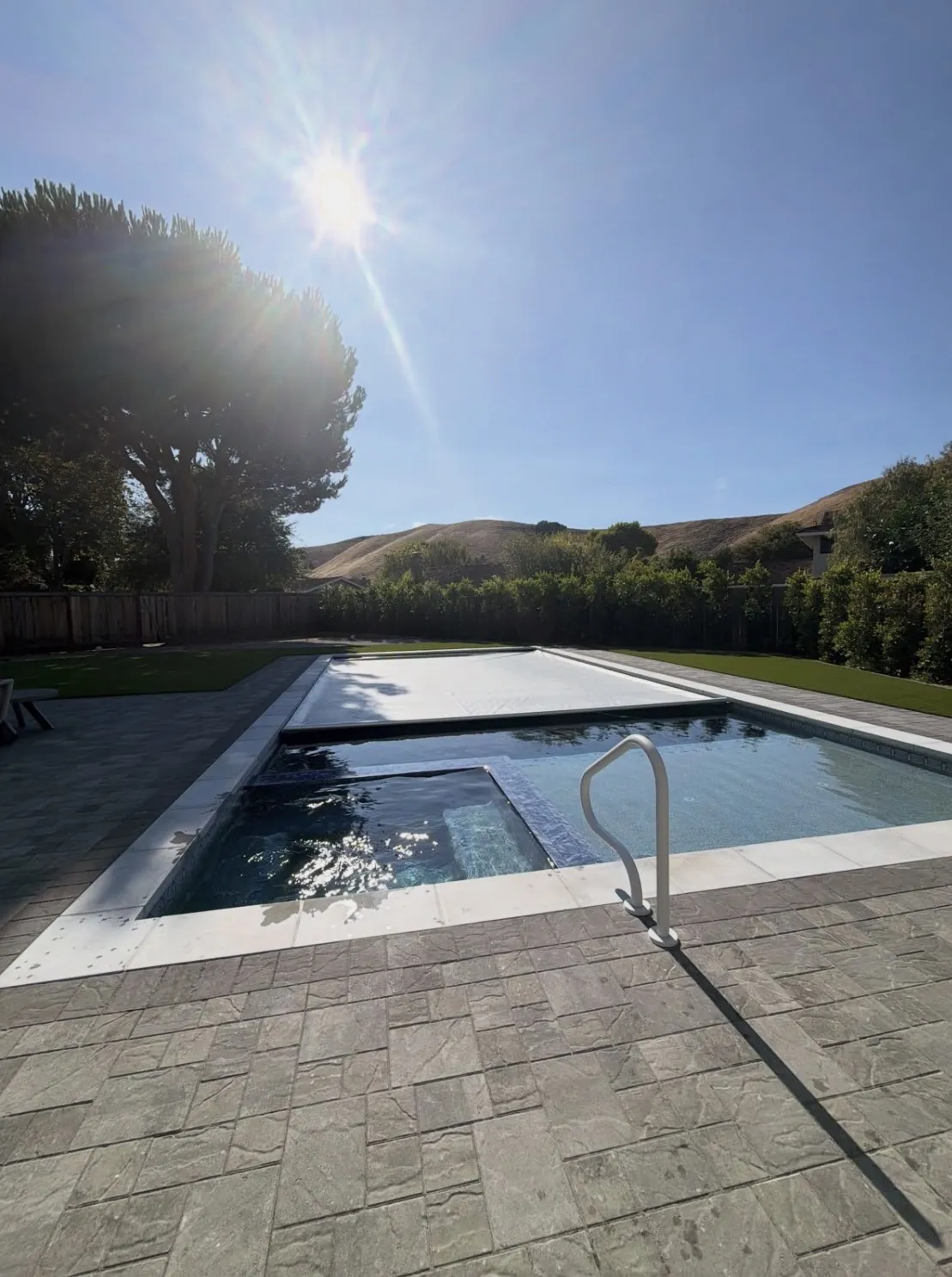Finished rectangular pool and integrated spa with mostly closed cover, surrounded by grey pavers and artificial turf.
