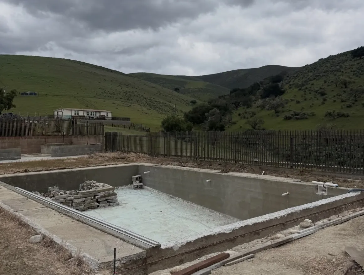Empty rectangular swimming pool shell under construction with a stack of stones inside and green hills in the background.