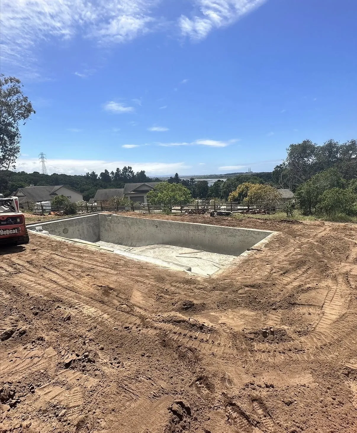 Empty rectangular gunite pool shell surrounded by bare dirt with heavy machinery tracks in a residential backyard.
