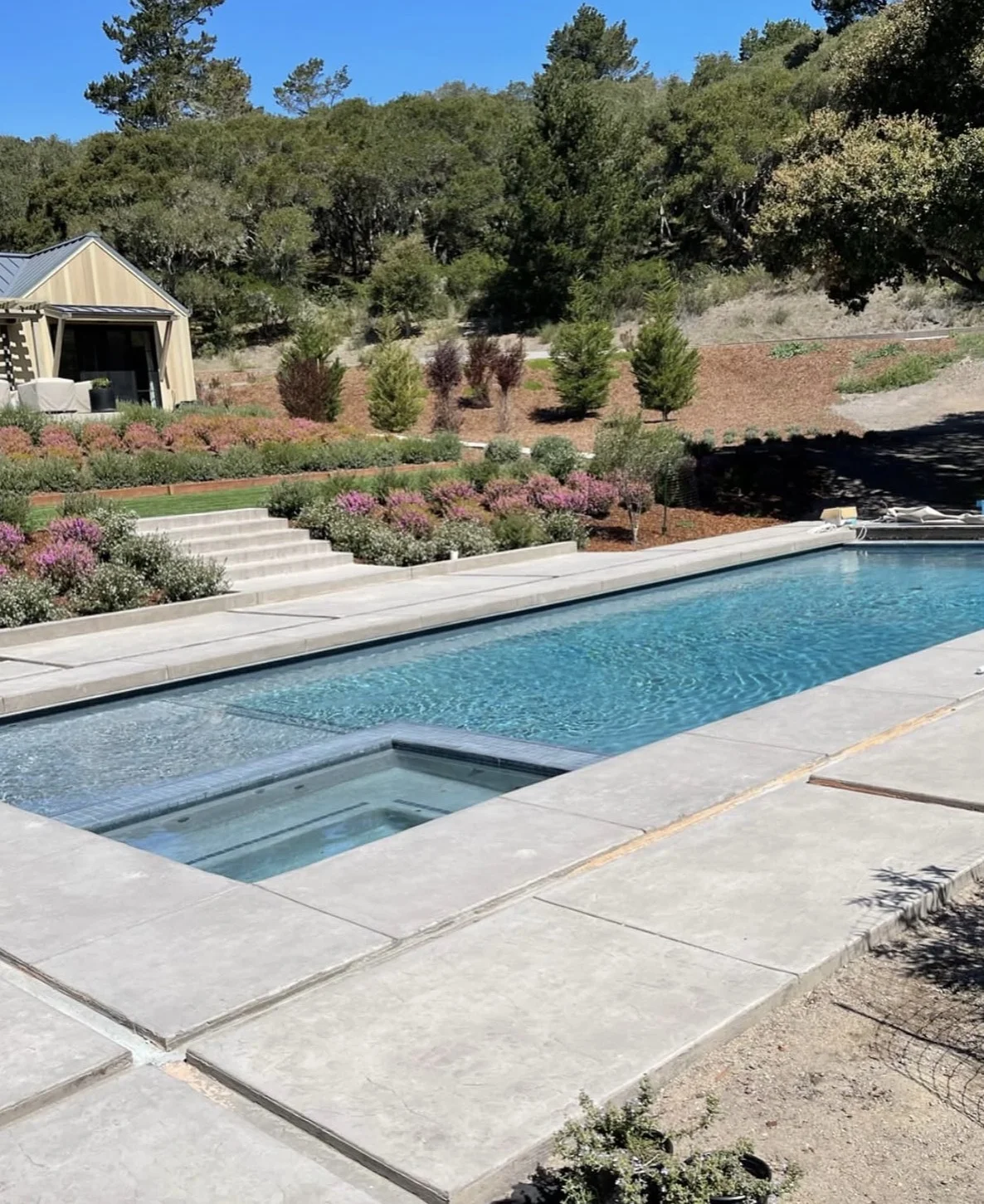 Finished rectangular swimming pool and integrated spa surrounded by concrete decking in a landscaped hillside backyard.