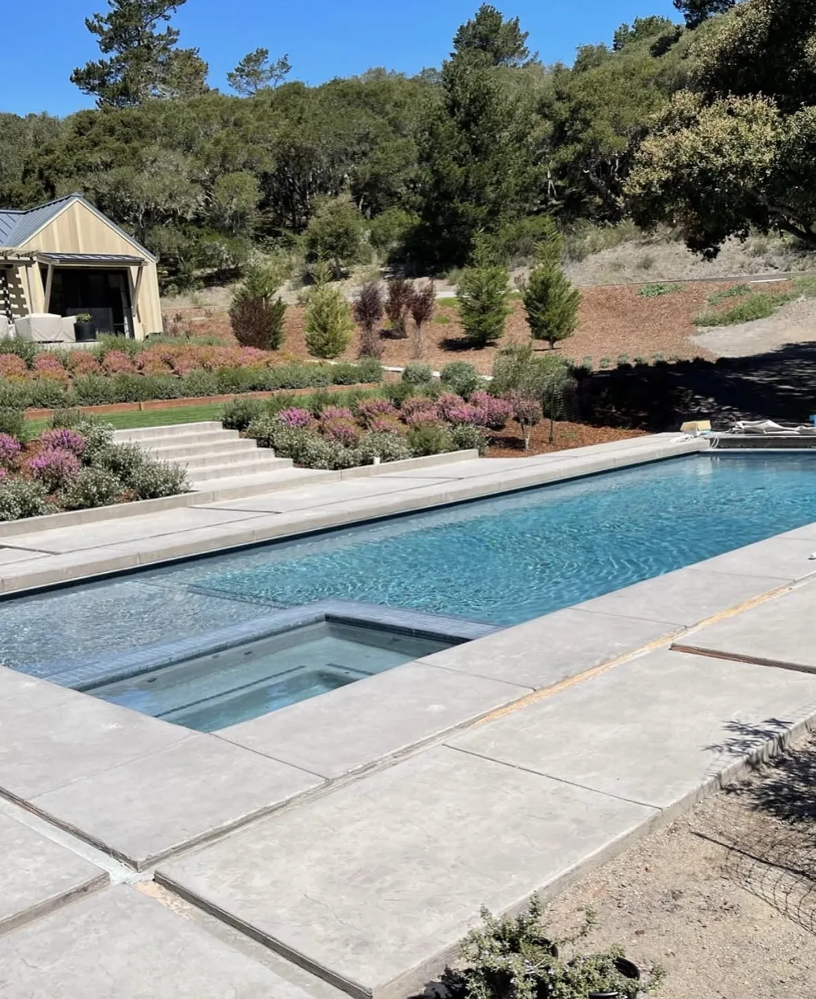 Finished rectangular swimming pool and integrated spa surrounded by concrete decking in a landscaped hillside backyard.