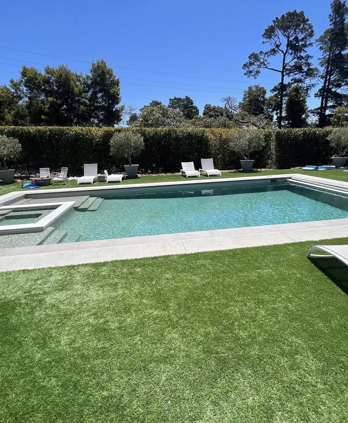 Rectangular swimming pool and integrated spa surrounded by artificial turf and white lounge chairs.