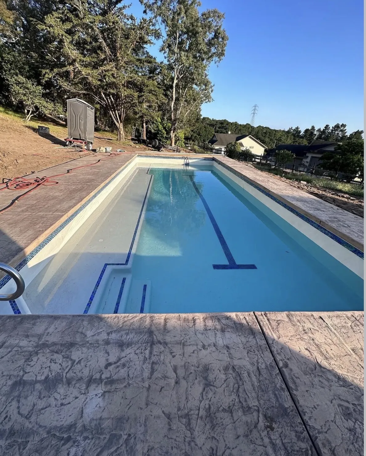 Rectangular swimming pool with a tanning ledge and blue tile lane line, surrounded by a stamped concrete deck and bare dirt.