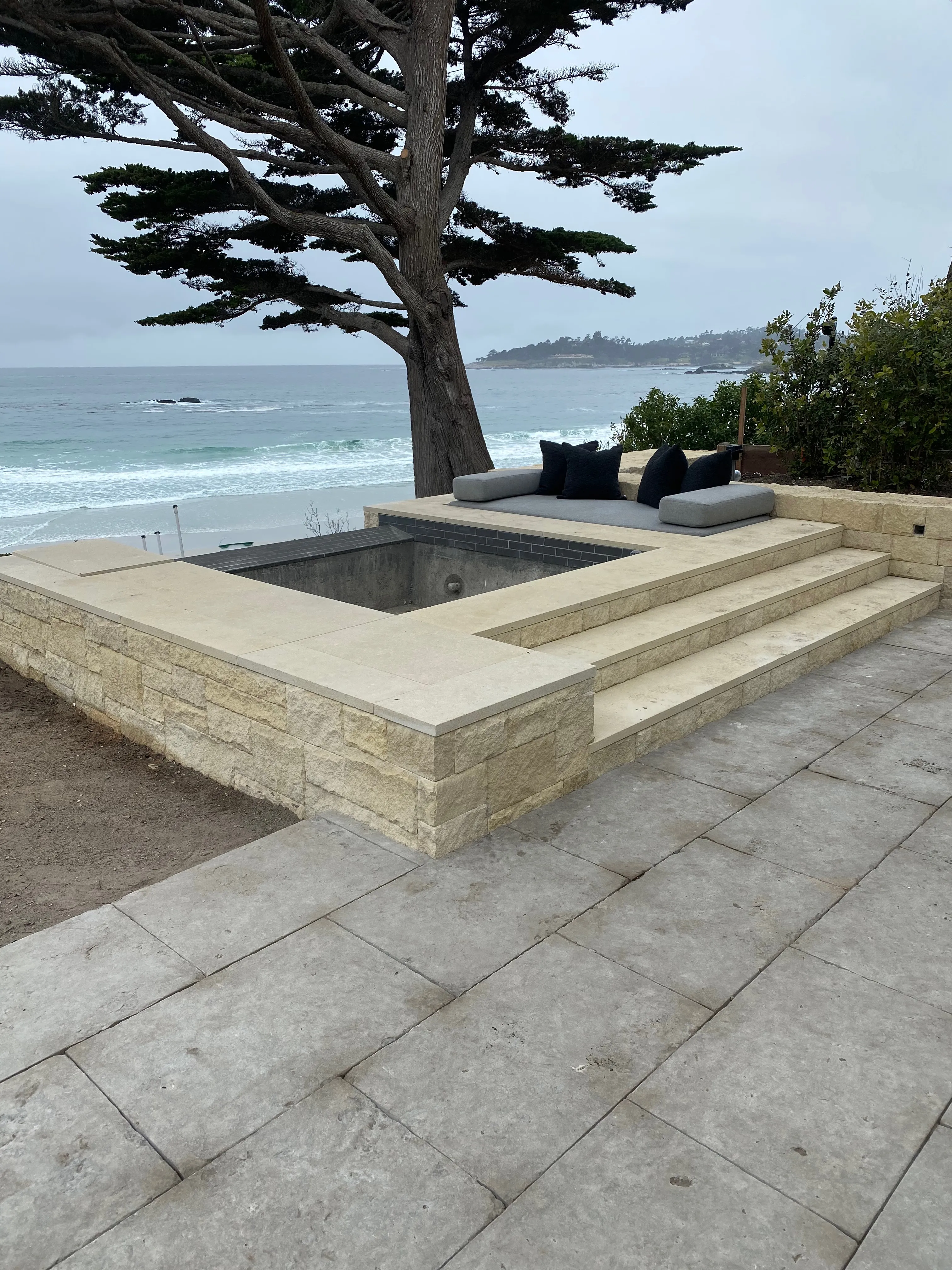 Built-in stone seating area with grey cushions overlooking the ocean next to a large pine tree.