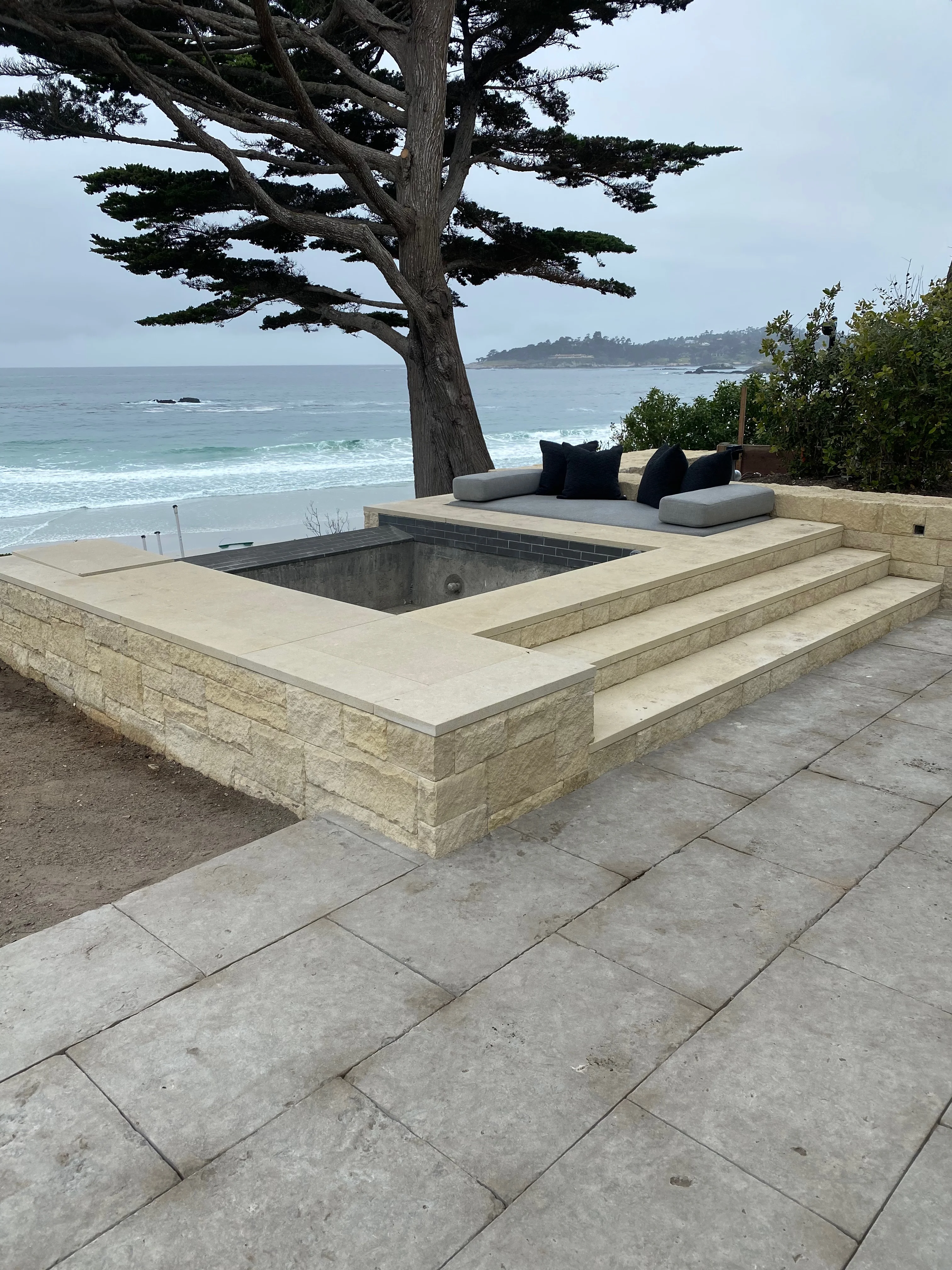 Built-in stone seating area with grey cushions overlooking the ocean next to a large pine tree.