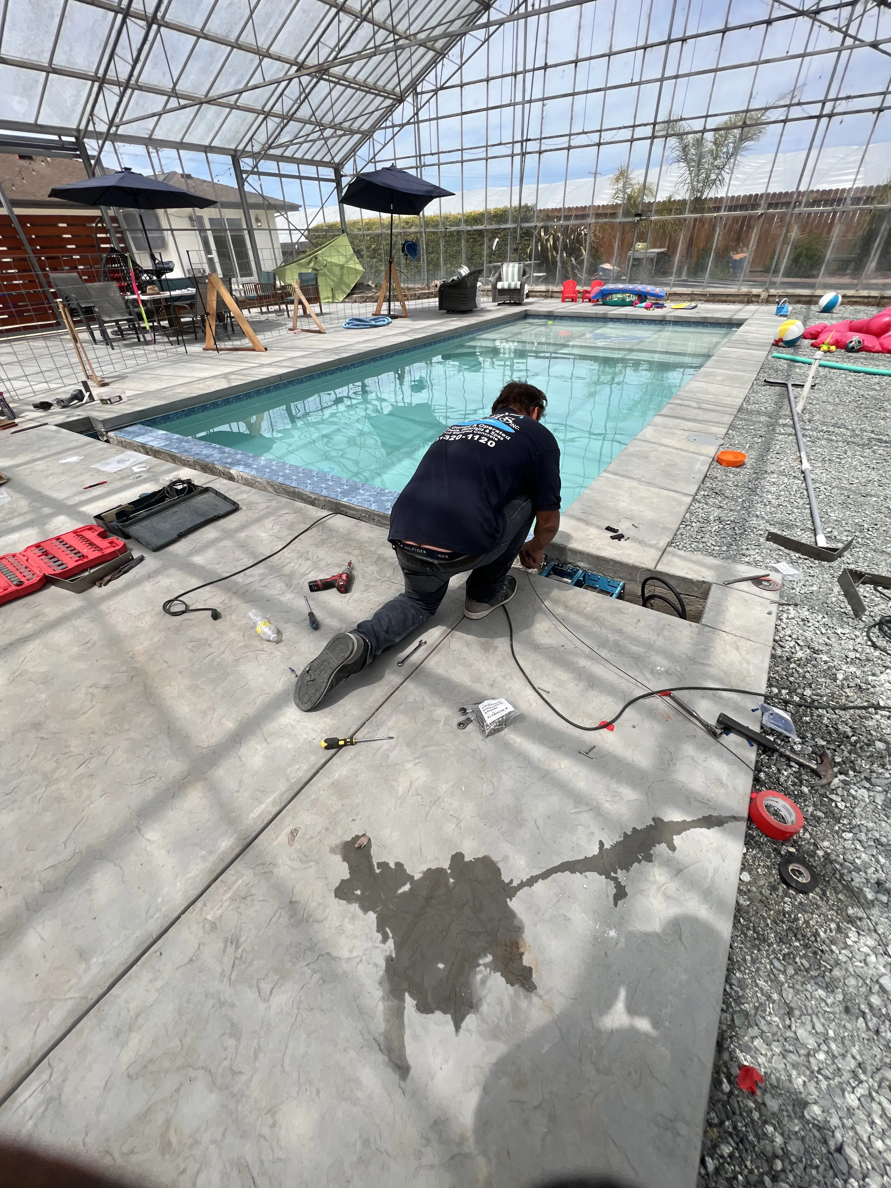 A worker kneels on a stamped concrete deck installing an automatic cover track alongside a filled pool.